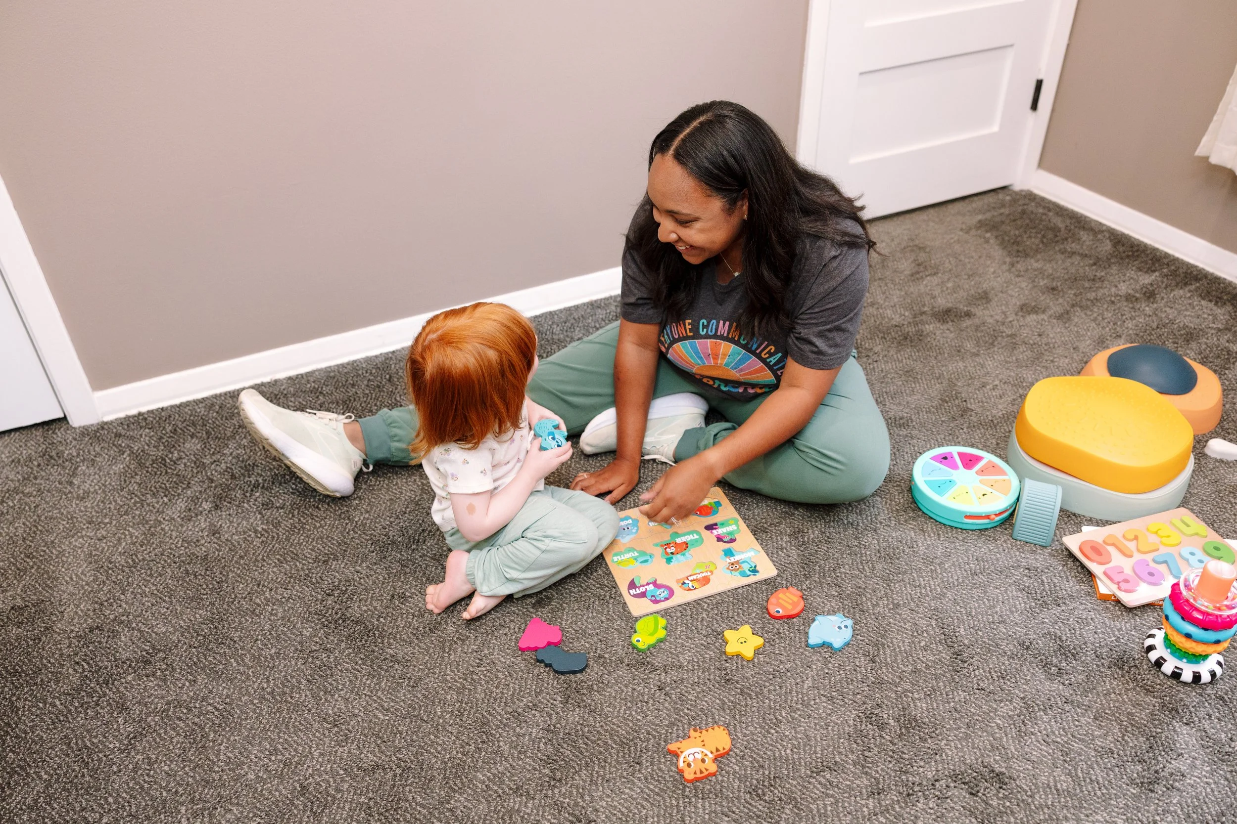 A woman and a young girl sitting on the carpeted floor playing with educational toys and puzzles, surrounded by colorful toys and games.