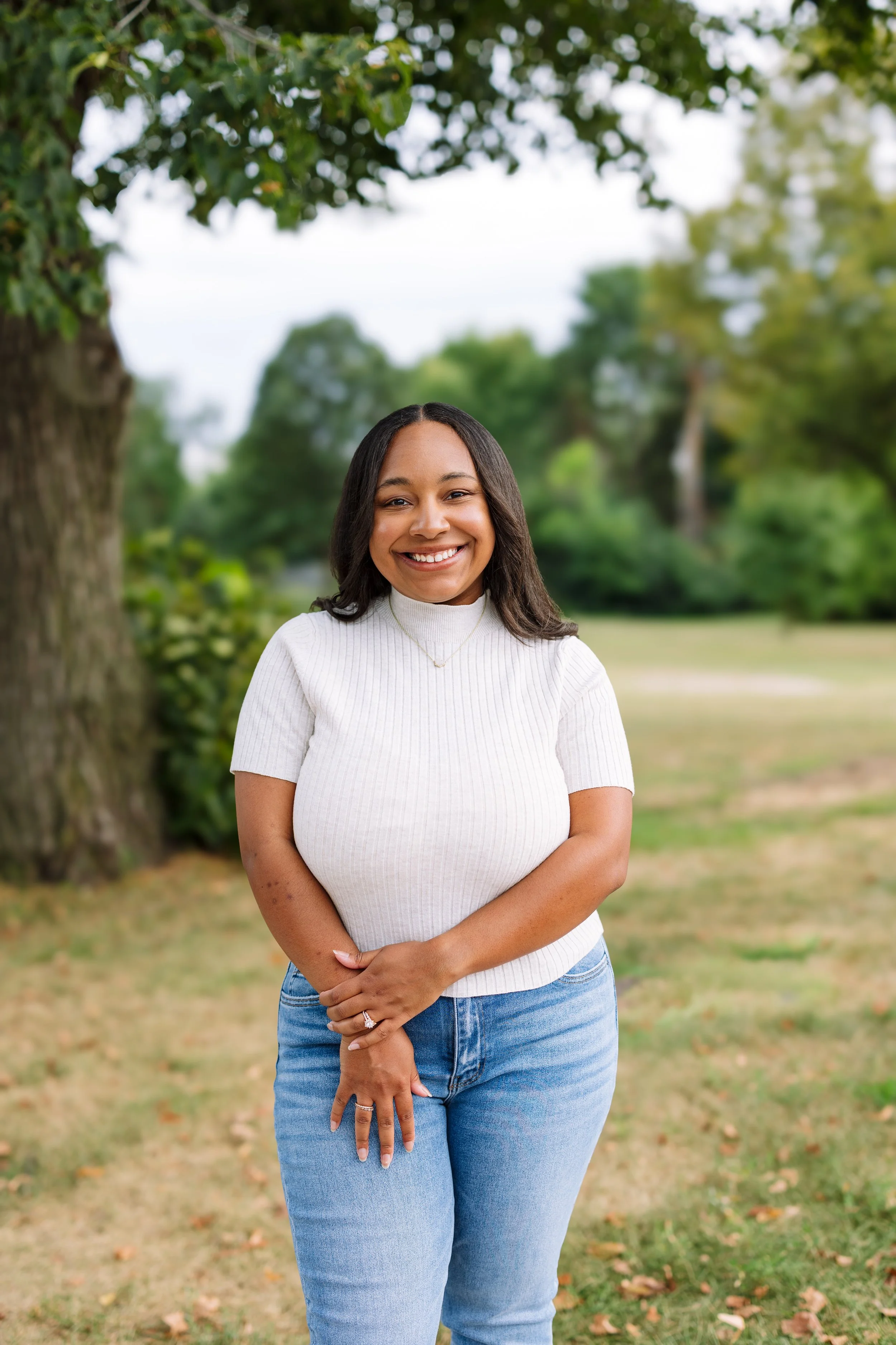 A young woman with dark hair smiling outdoors near a large tree and green foliage, wearing a white ribbed turtleneck sweater and blue jeans.