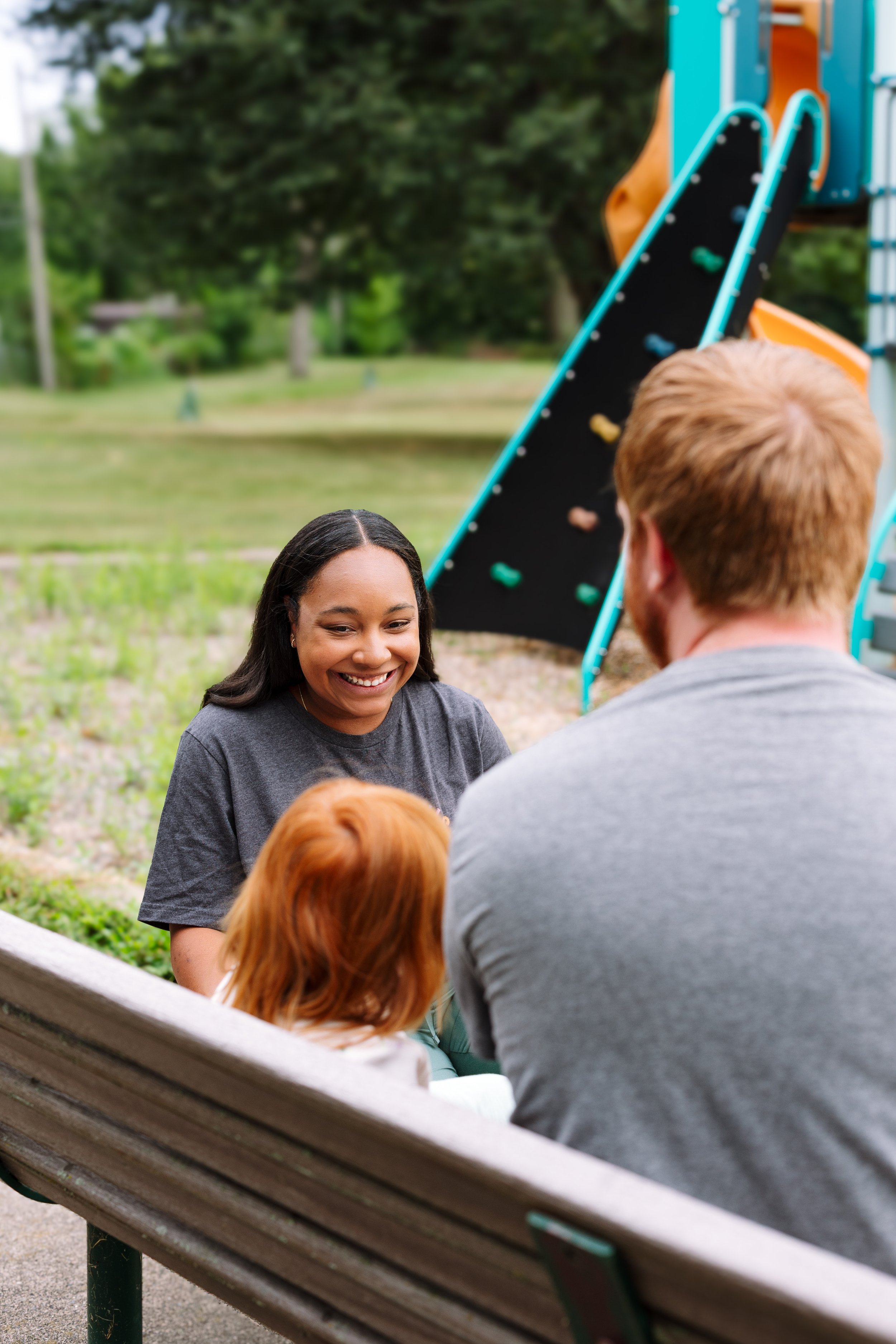 A young woman smiling and talking to a man on a park bench, with a playground slide and green trees in the background.