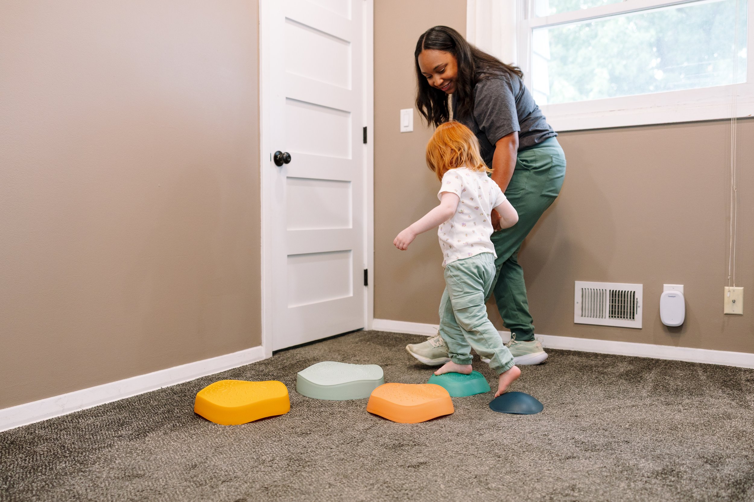 A woman and a child playing on a set of colorful stepping stones inside a home, near a window and door.