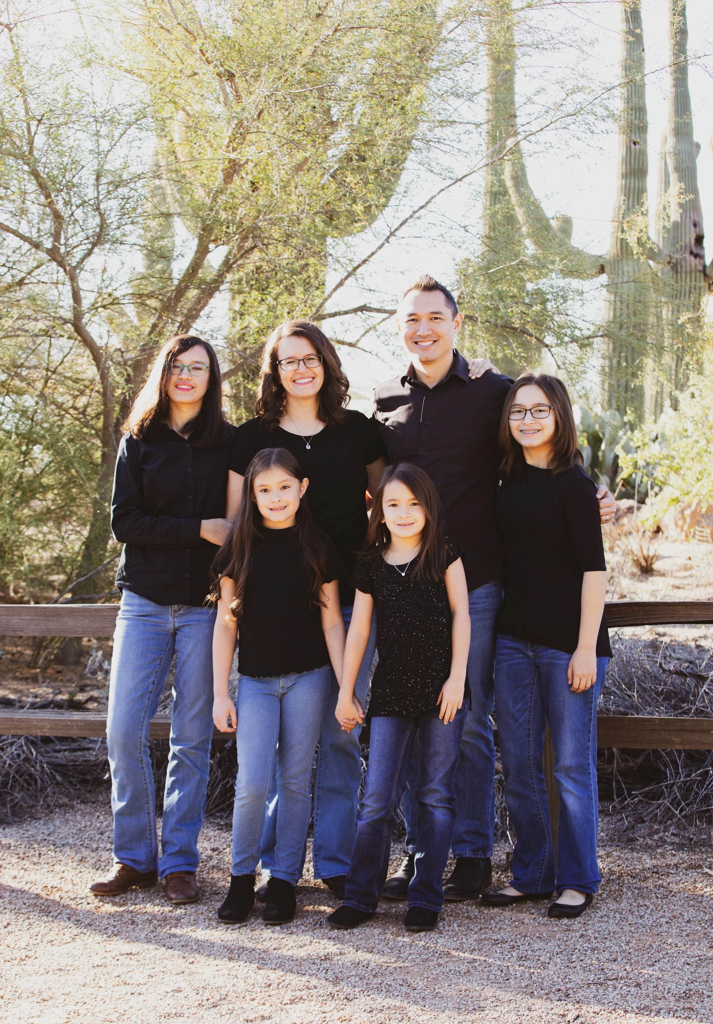 A group of seven people standing outdoors on a sunny day, with five women and two children in the front and two men in the back, all smiling and wearing casual black tops and jeans.