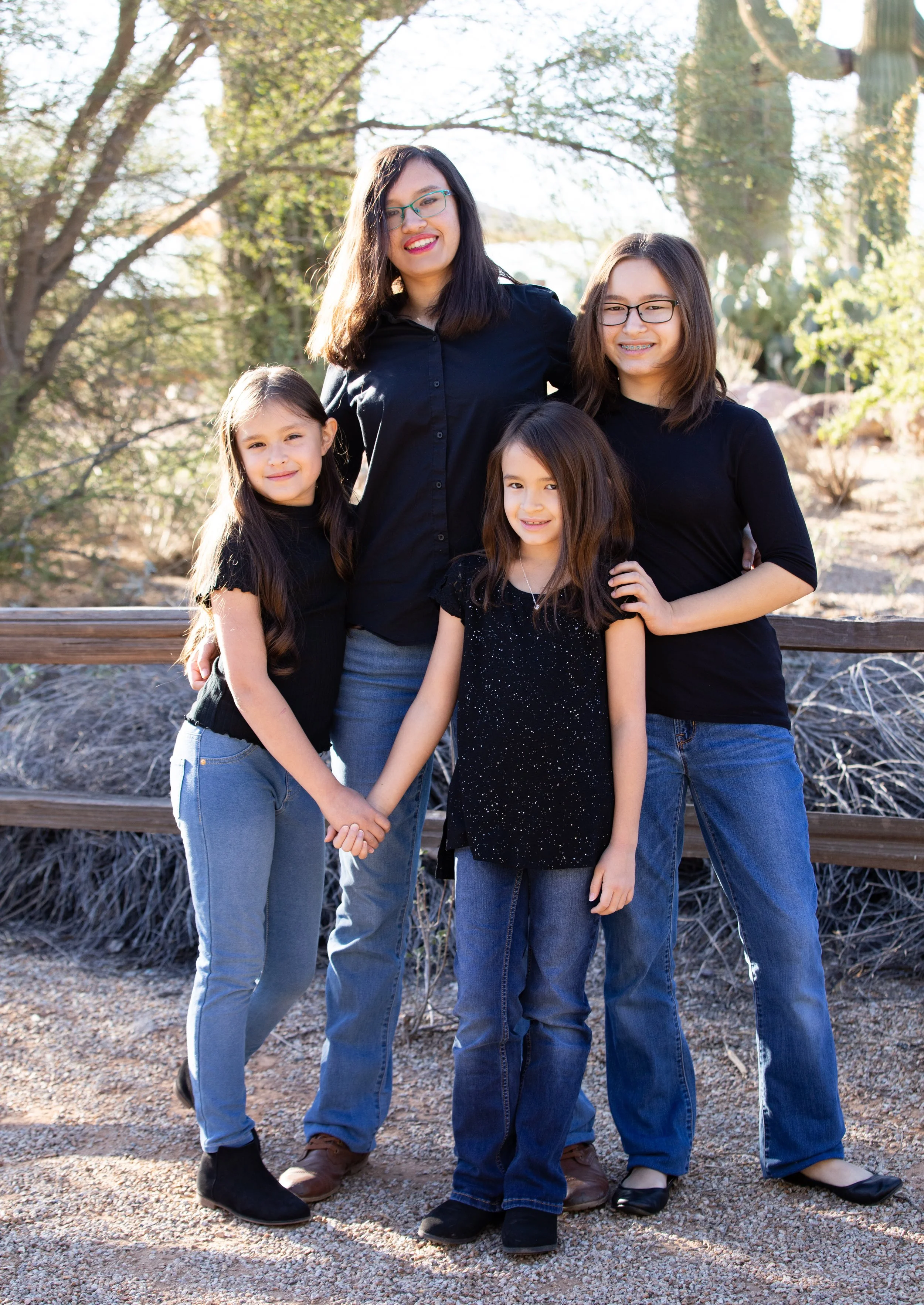 Four females, including one adult and three children, standing outdoors in front of trees, smiling, and holding hands.