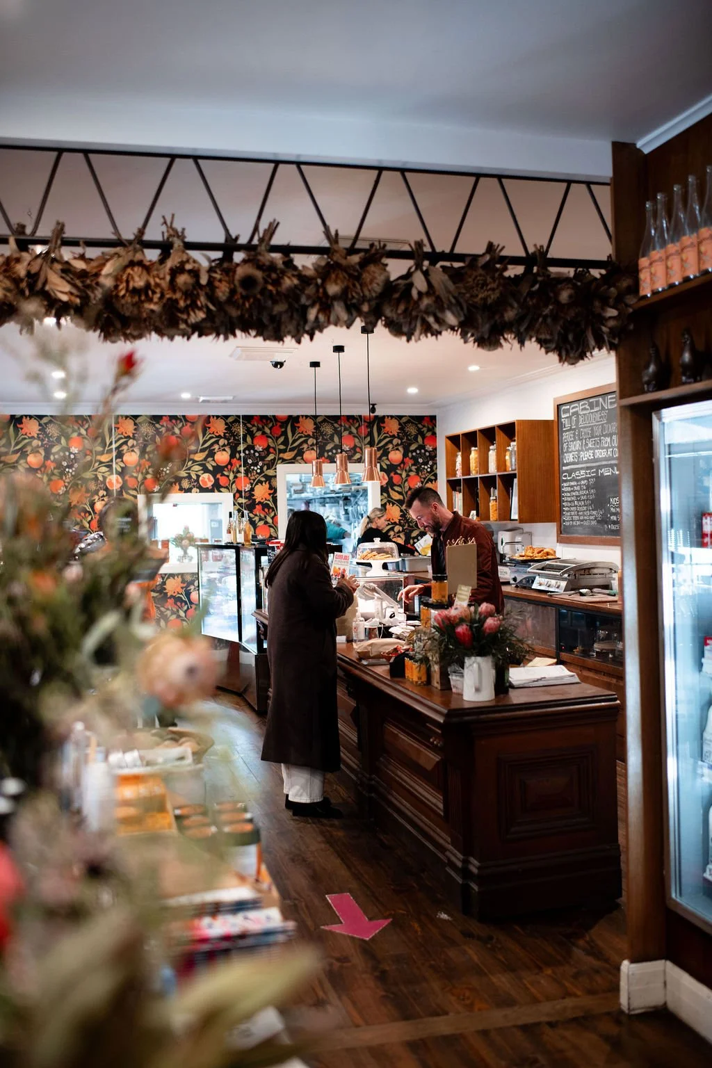 Interior of a cozy cafe with a floral wallpaper, a decorated counter, and customers ordering. Dried flowers hang from the ceiling.