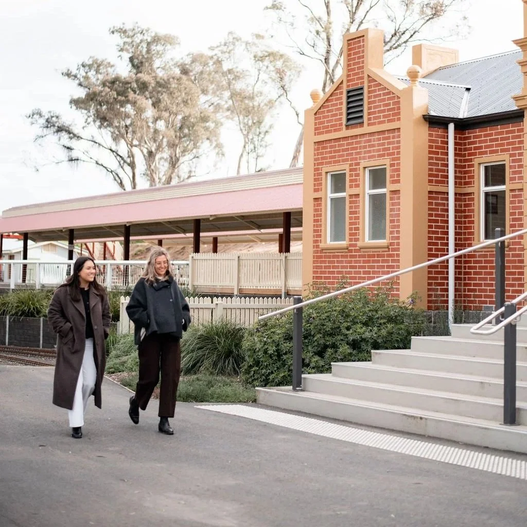 Two women walking and talking outside a brick building with stairs, handrail, and a covered area in the background, trees beyond.