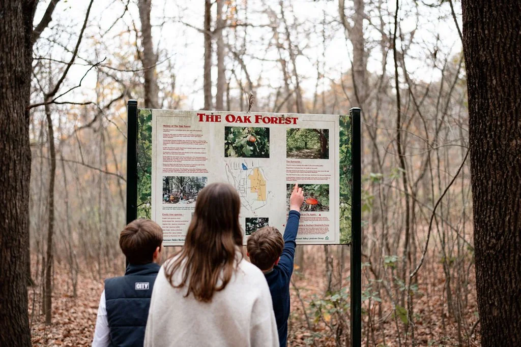 Three children reading an informational sign about the Oak Forest in a wooded area during autumn.