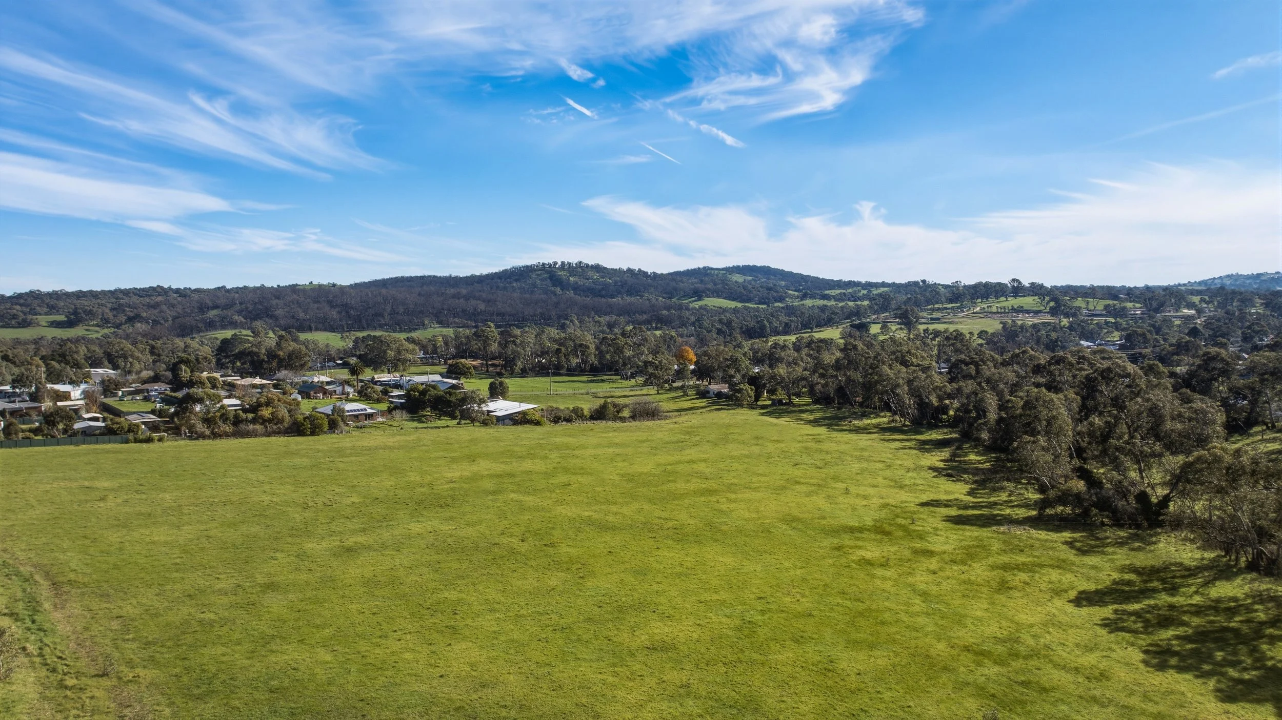 A scenic landscape of rolling green hills, scattered trees, and a small residential area beneath a bright blue sky with wispy white clouds.