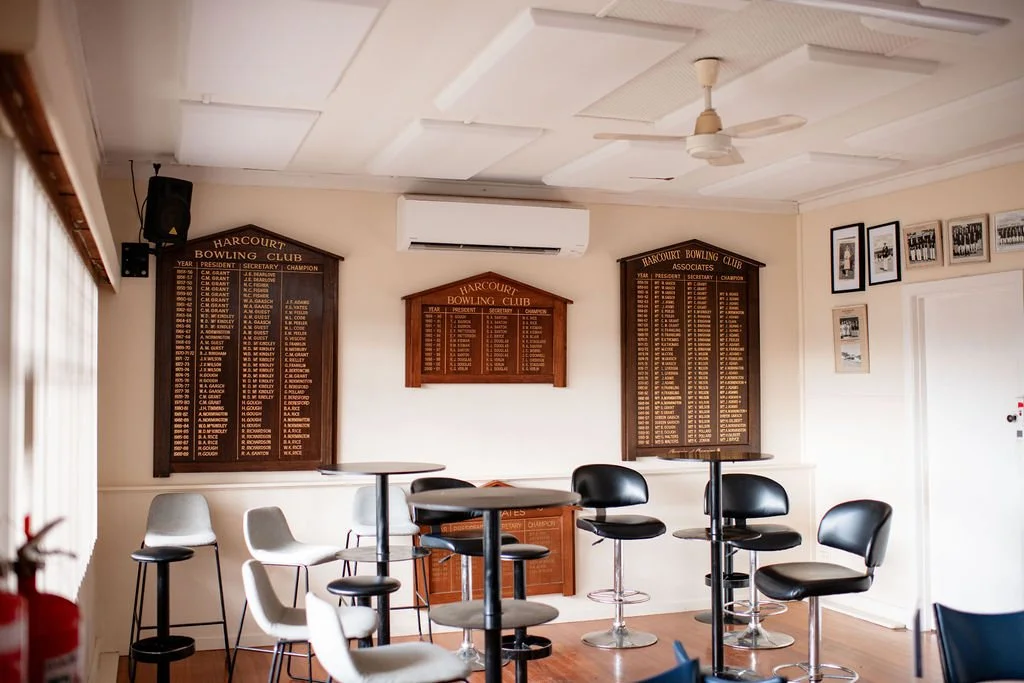 Interior of a bowling club lounge with wooden plaques displaying hall of fame names and awards, tables and chairs, framed photos on the wall, an air conditioning unit, and ceiling fan.