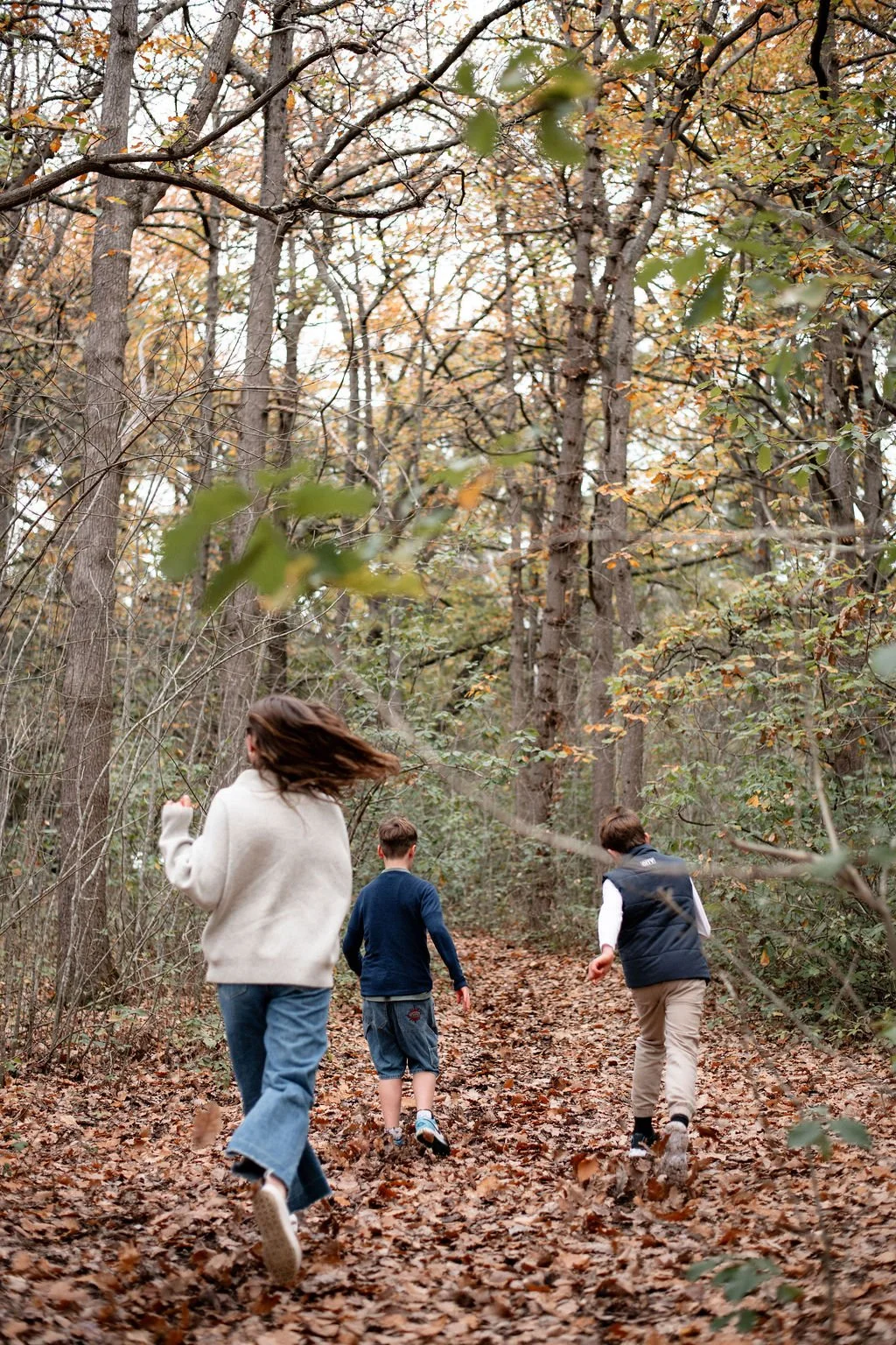 Three children running on a leaf-covered trail in a forest during autumn.