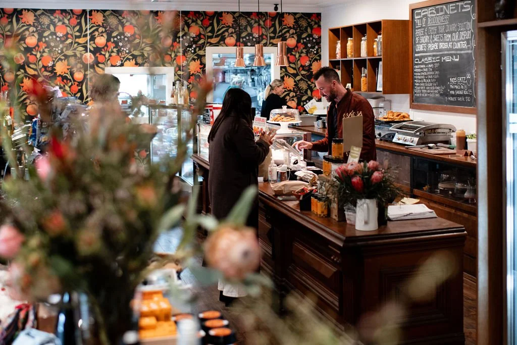 Customers ordering at a cafe counter with a floral wallpaper background and a chalkboard menu.
