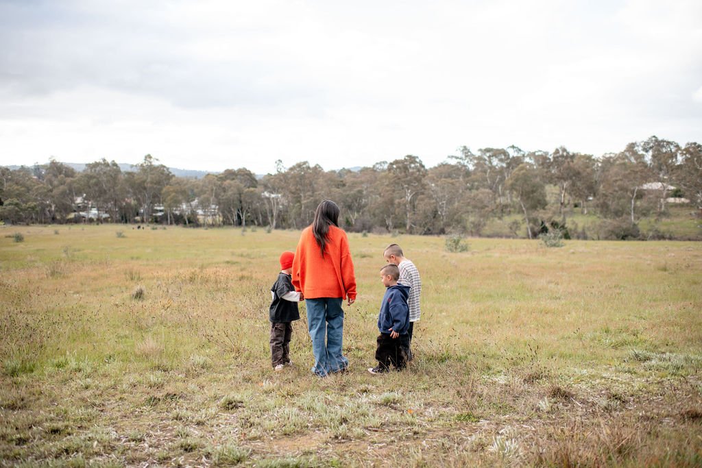 A woman and four children standing together in a grassy field on a cloudy day, with trees in the background.