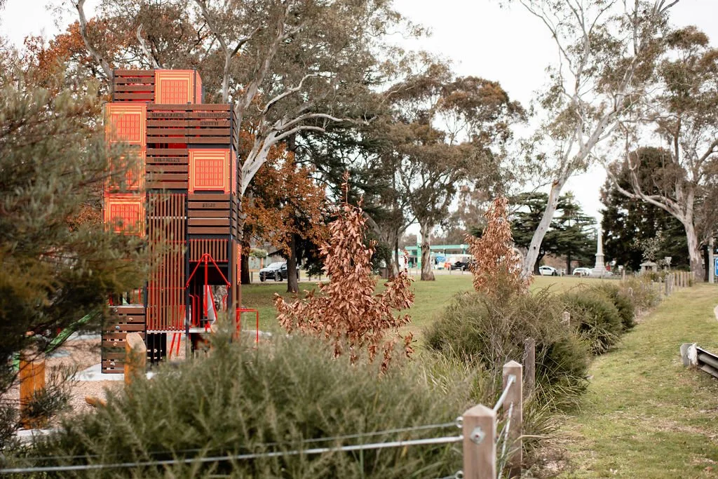 A park scene with trees, bushes, and a modern wooden playground structure on the left, and a grassy pathway with a white fence on the right. In the background, there are cars and a small building.