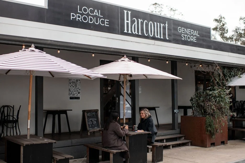 Two women sitting outside at a small table under striped umbrellas, in front of a store called Harcourt, which sells local produce and functions as a general store.