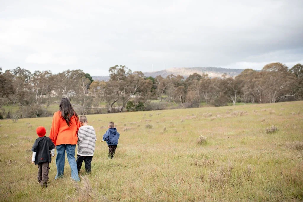 A woman and four children walking in a grassy field with trees and distant hills in the background under a cloudy sky.