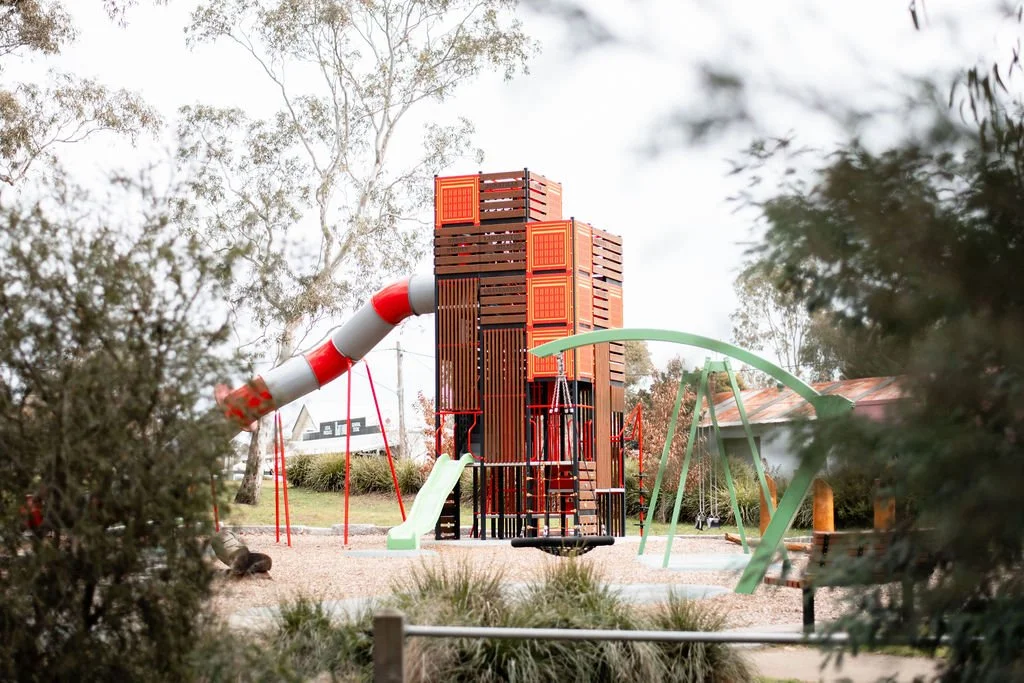 A playground with a large wooden tower structure, slides, and swings, surrounded by trees and natural scenery.