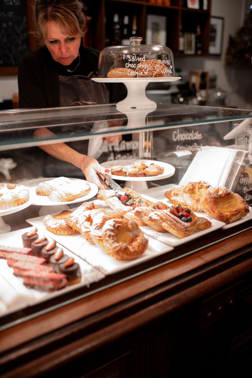 A woman behind a glass display case filled with assorted pastries, including croissants, Danish pastries, and fruit-topped treats, in a bakery or cafe setting.