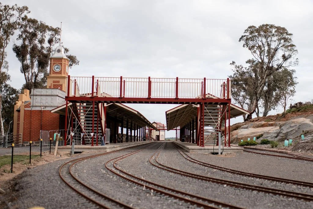 A small train station with train tracks, a clock tower, and an overpass in a rural setting.