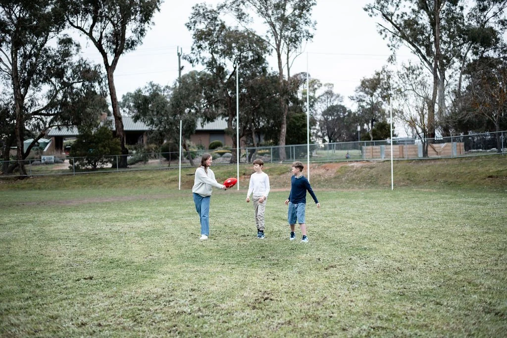 A woman holding a football coaching two boys on a grassy field outdoors, with trees and a fence in the background.