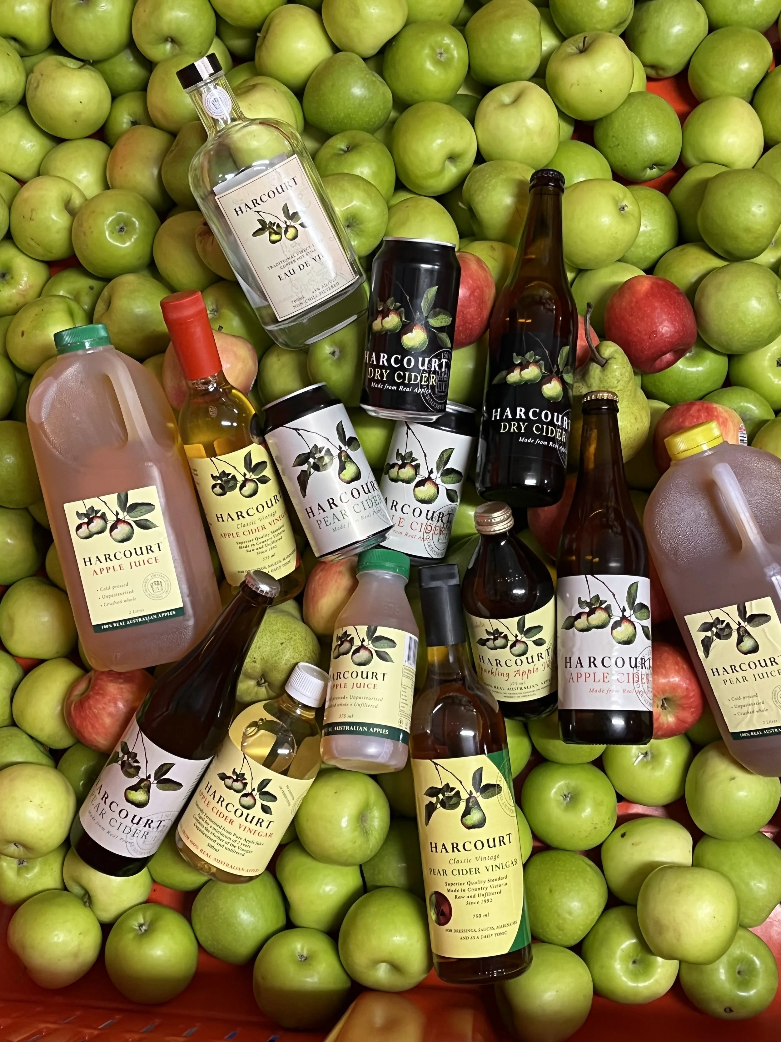A basket filled with green apples and red apples, with several bottles of Harcourt apple cider, apple juice, pear cider vinegar, and dry cider scattered on top of the apples.