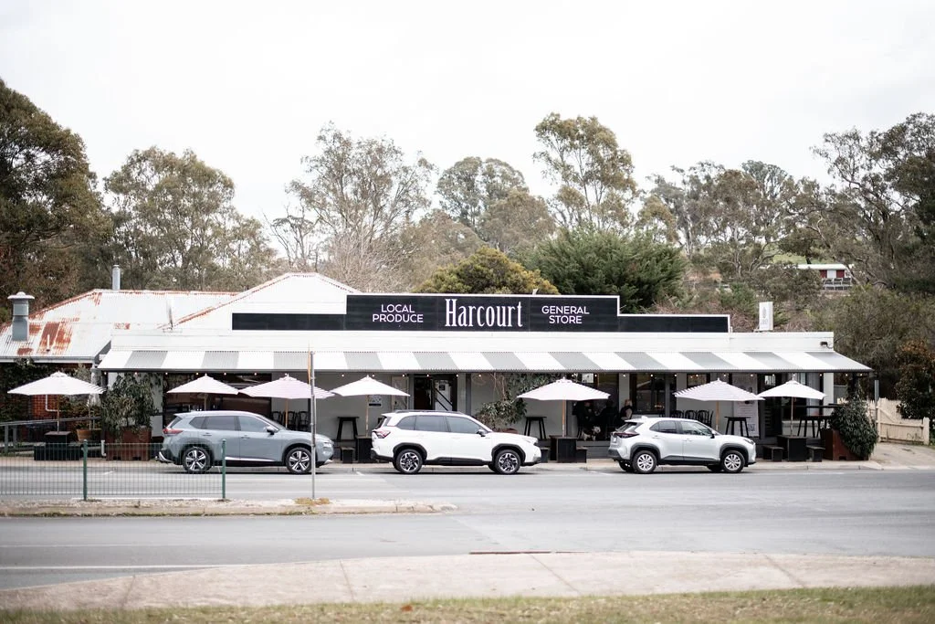 Exterior of Harcourts store with cars parked in front and outdoor seating with umbrellas.