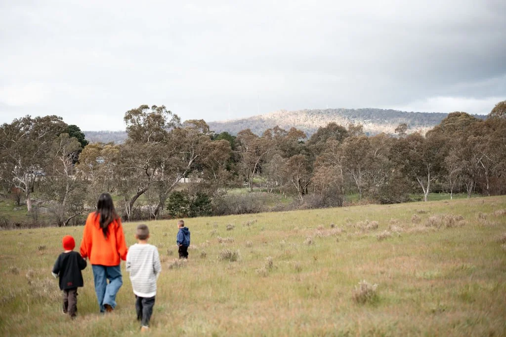 Four people, including three children and an adult, walking in an open grassy field with trees in the background under a cloudy sky.