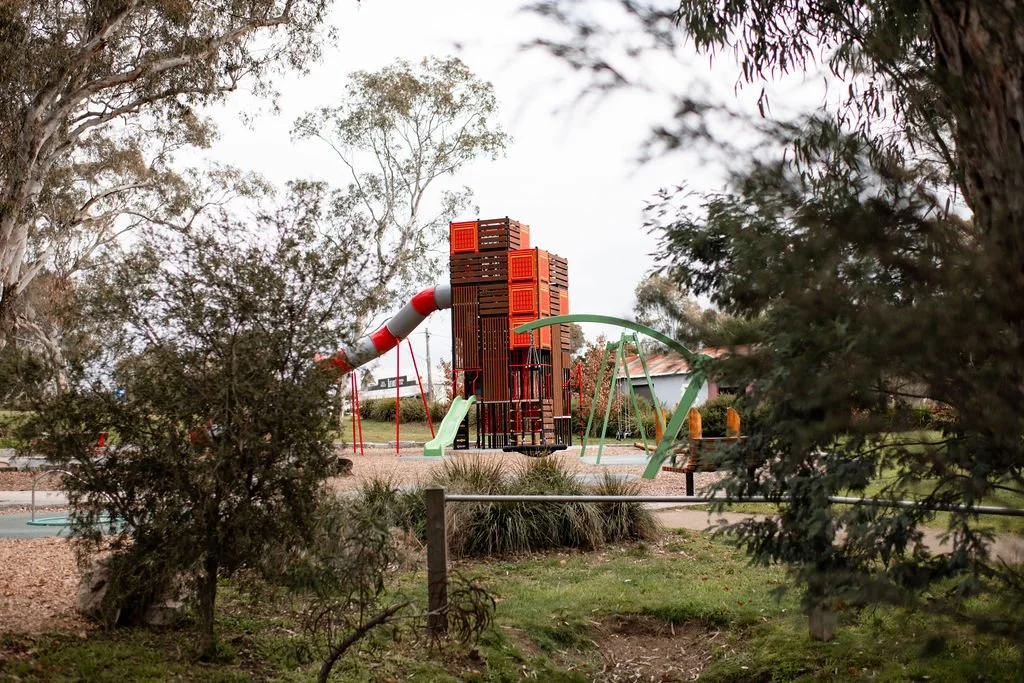 A playground with a large play structure including slides, a tunnel, and climbing elements, surrounded by trees and greenery.