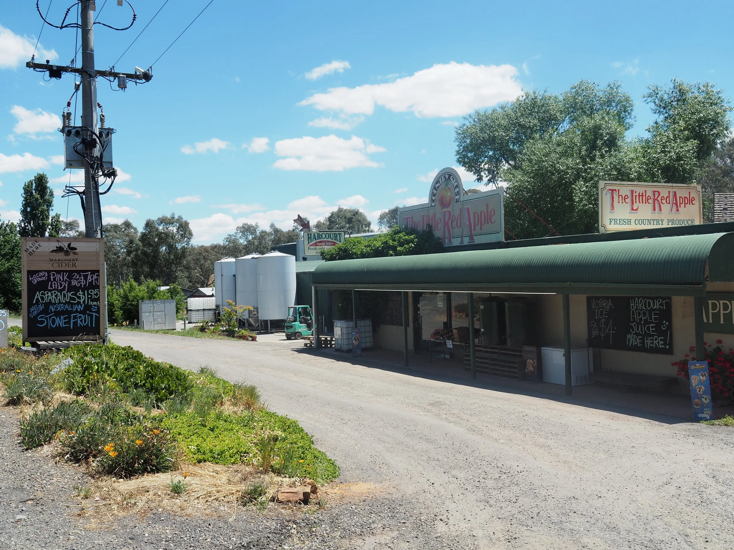 A roadside market with signs advertising fresh local produce and apple juice, and a chalkboard menu listing items like lady bags and stone fruit. There is a utility pole in the foreground and trees in the background under a partly cloudy blue sky.