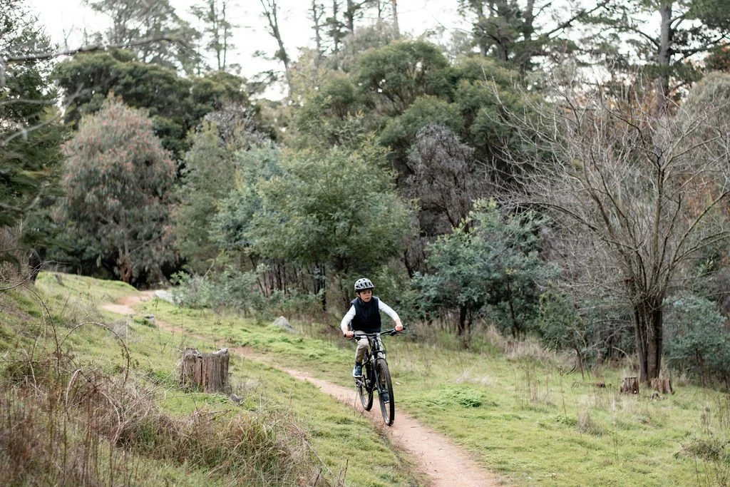 Child riding a mountain bike on a dirt trail through a wooded area with trees and shrubs.