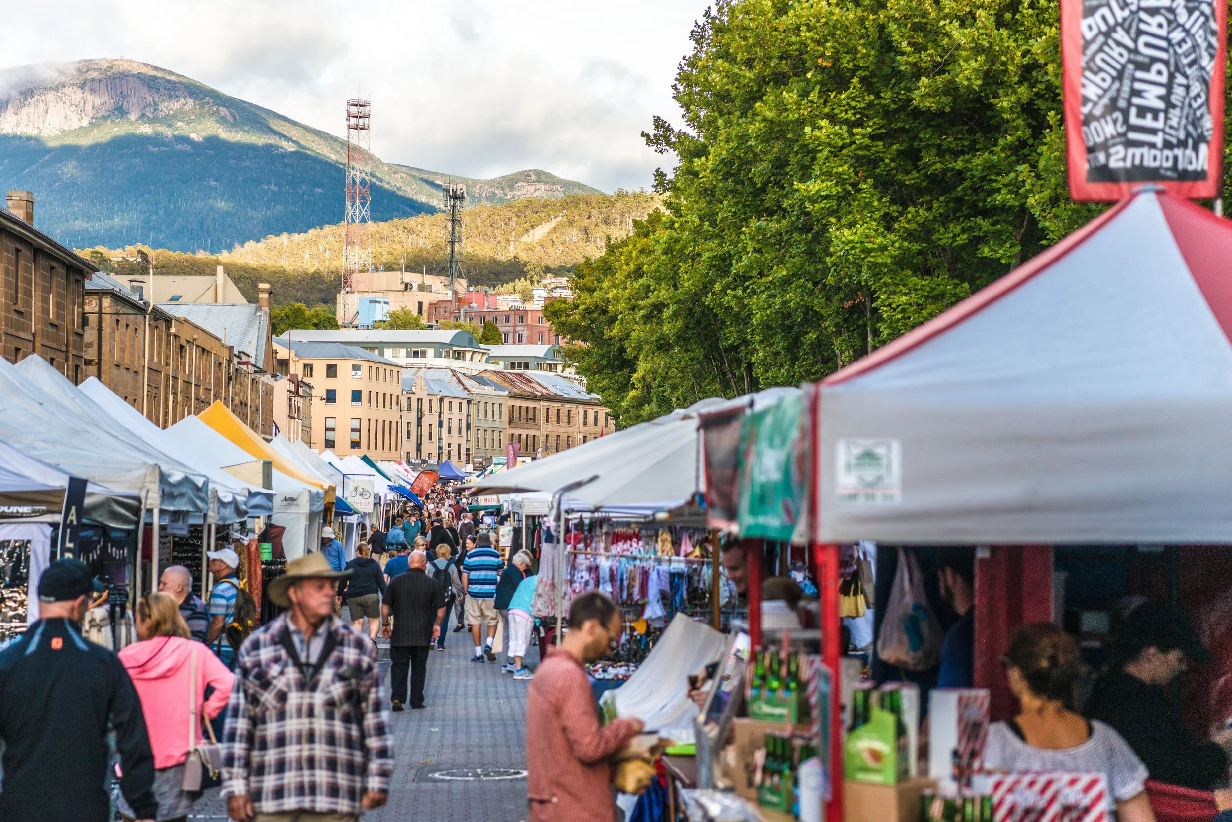 Salamanca Market, a busy outdoor market with numerous tents and stalls, filled with people shopping and walking. Tall buildings are behind the stalls, and a mountain with green forests and some clouds is in the background.