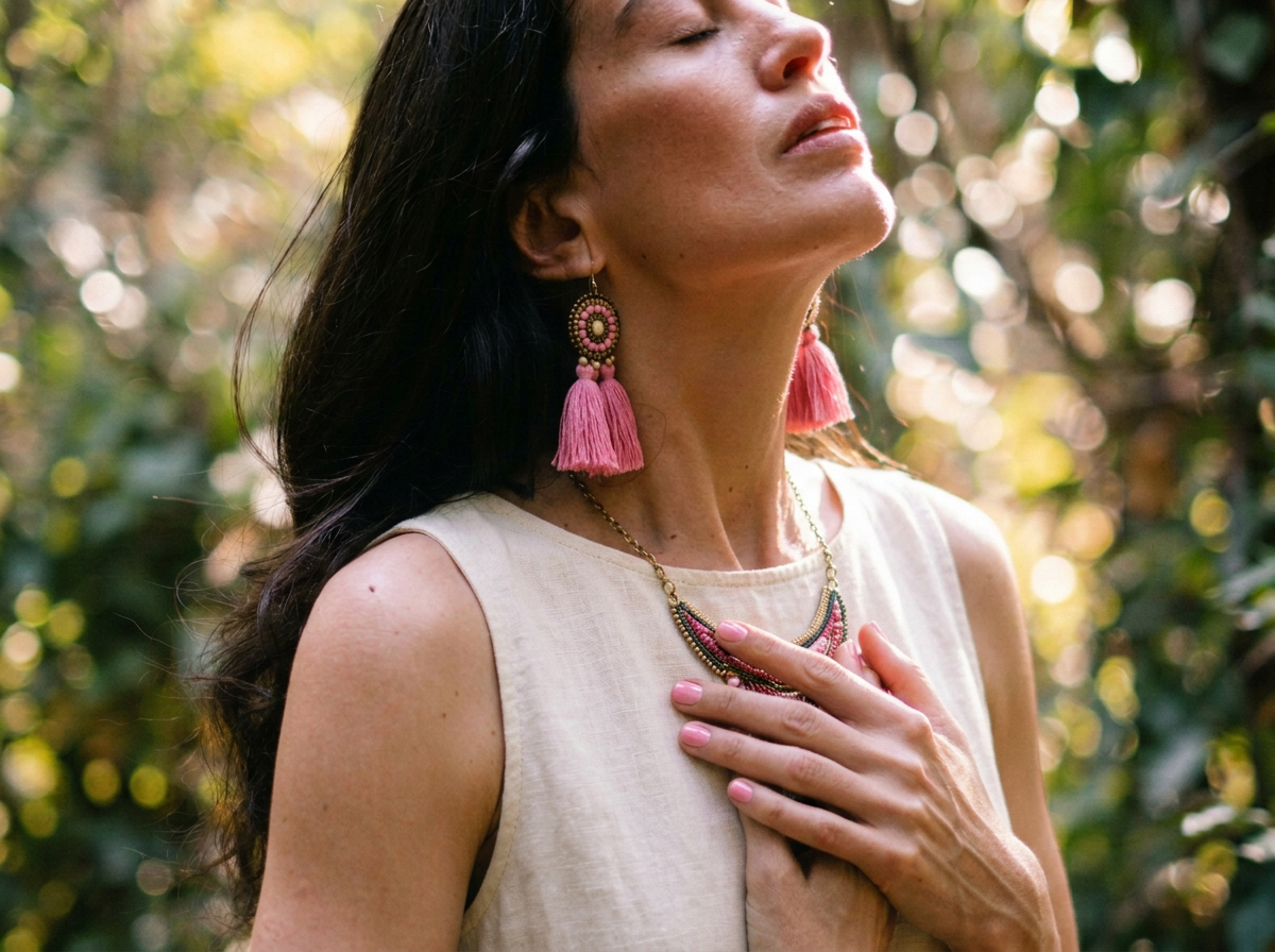 A woman with dark hair wearing pink tassel earrings, a beaded necklace, and a sleeveless cream top, standing outdoors with a blurred green background, her eyes closed and one hand over her chest.