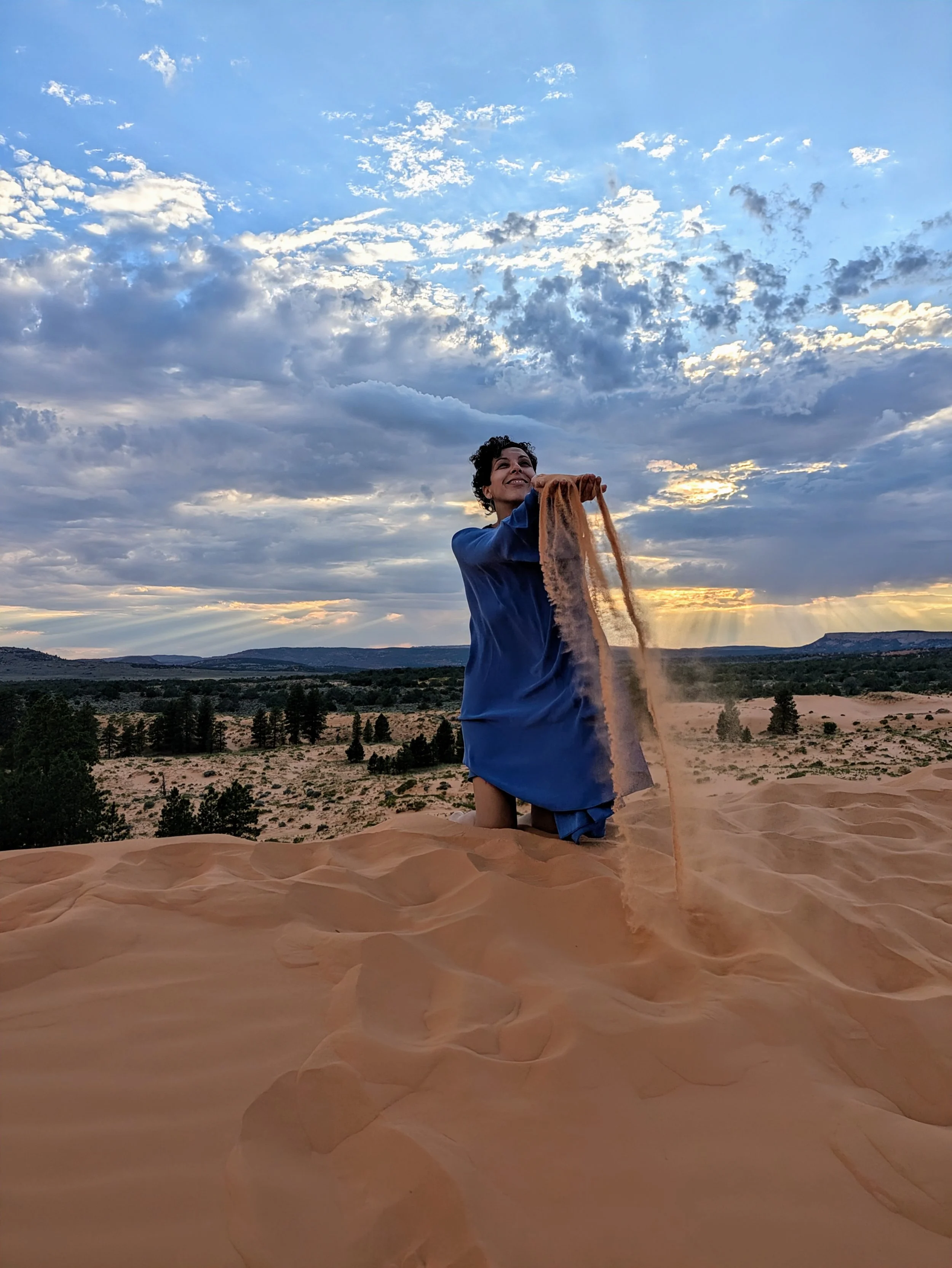 Woman kneeling on sand dune at sunset, holding sand flowing from her hand with a desert landscape and cloudy sky in the background.
