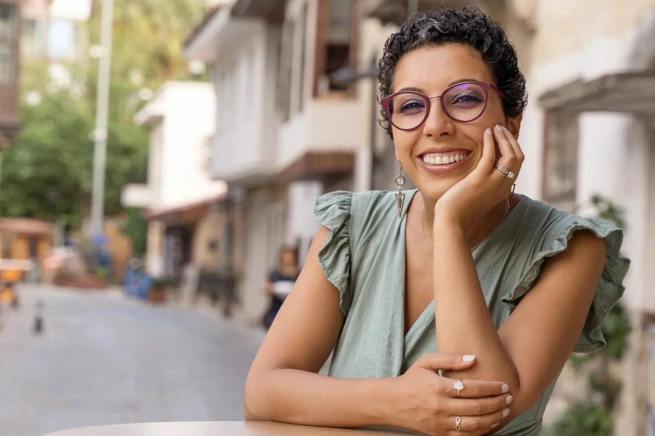 A woman with short curly dark hair wearing glasses with maroon frames, a light green top with ruffled sleeves, and jewelry, smiling in an outdoor urban setting.