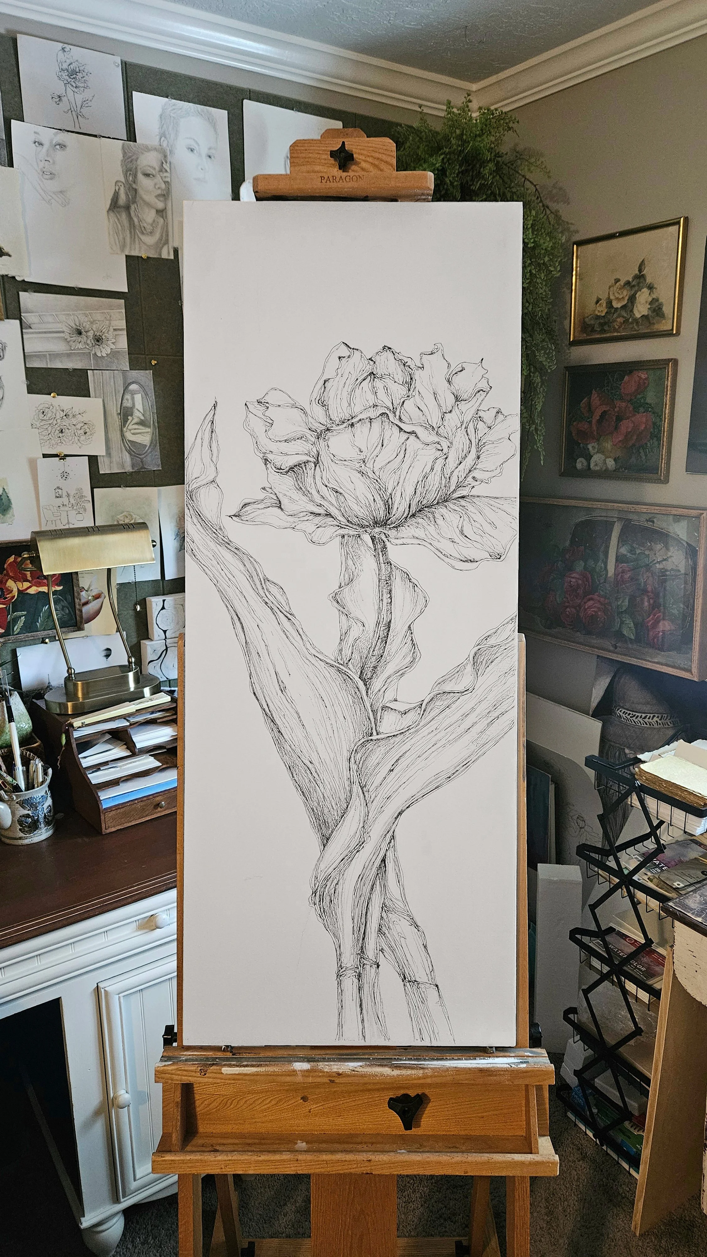 A large vertical canvas features a black and white line drawing of a flower, possibly an iris, with detailed petals and leaves. The canvas is on an easel in an art studio or gallery, surrounded by various artwork and supplies.