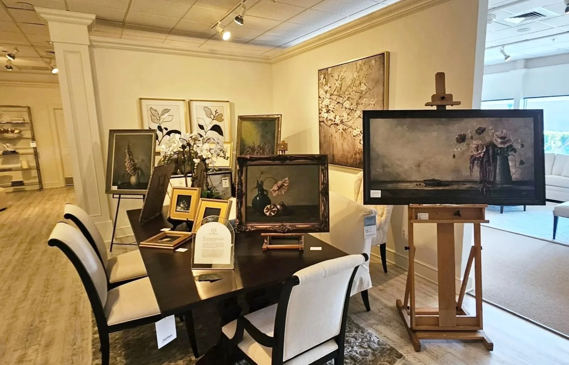 An art gallery displaying various framed paintings and a floral arrangement on a black table, with additional artwork hung on the cream-colored walls.