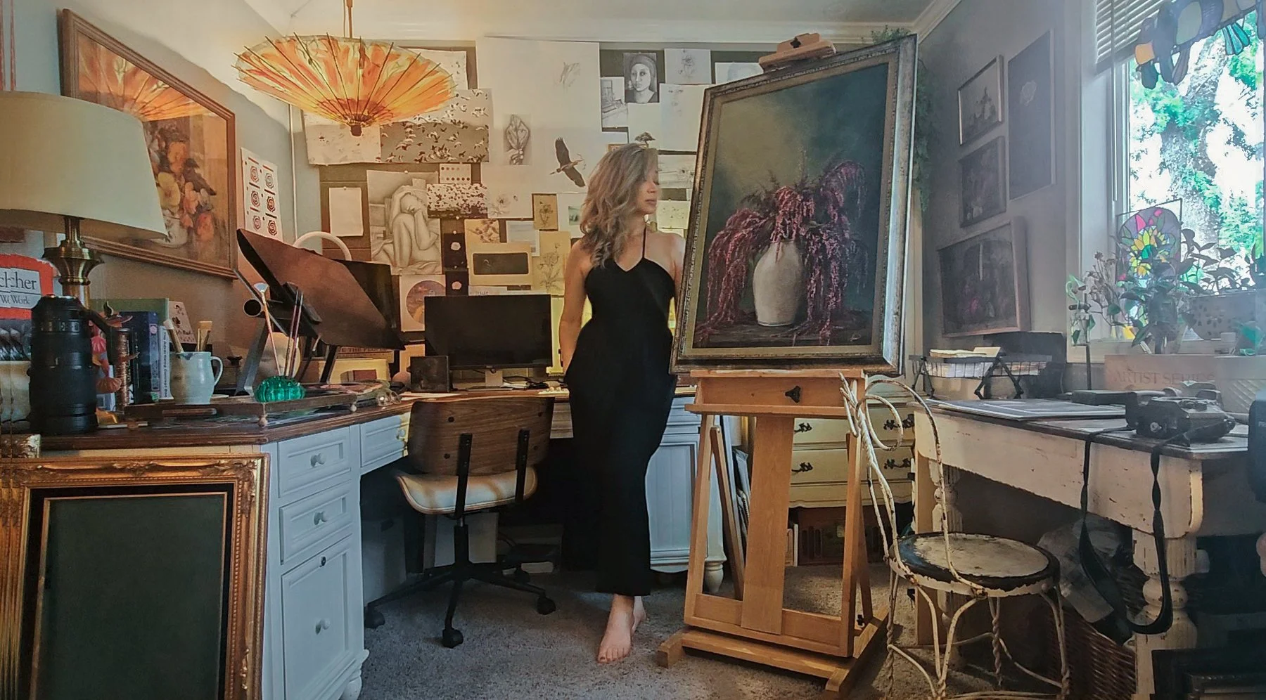A woman in a black dress stands barefoot in an art studio, looking at a large framed oil painting of a vase with flowers on an easel. The room is filled with various artworks hung on the walls, including sketches, paintings, and photographs, as well as art supplies and furniture.