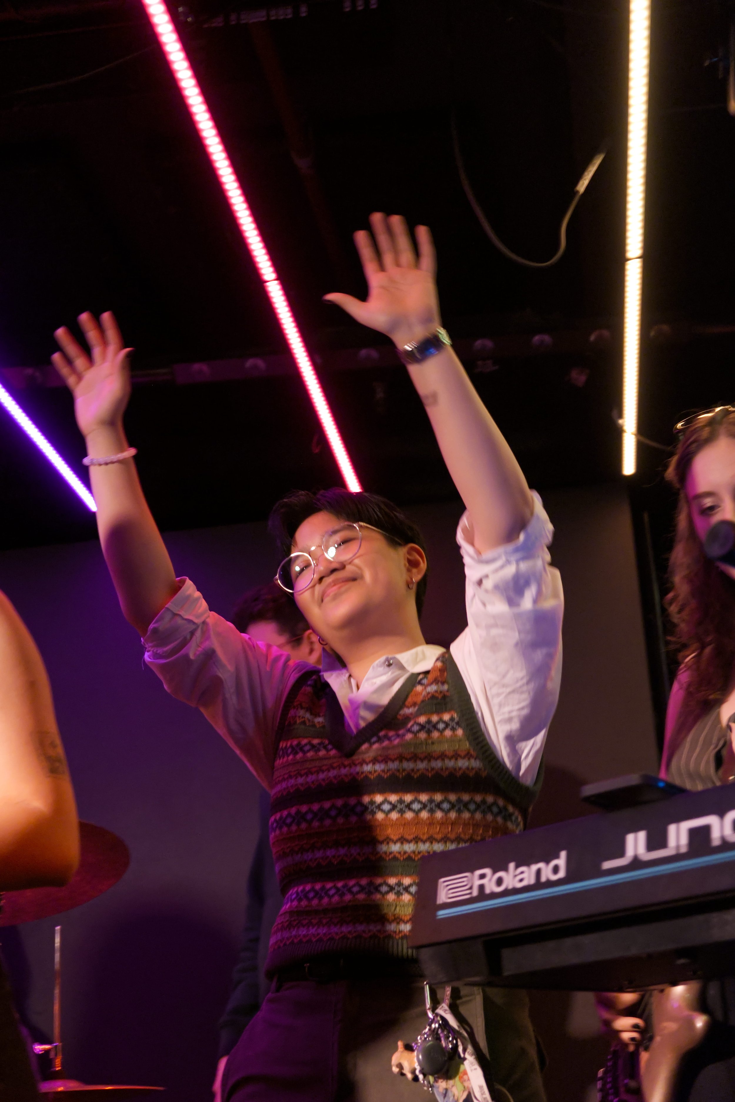 A person with glasses is standing with their arms raised, demonstrating excitement or celebration. The background features colorful LED strip lights, and part of a Roland electronic musical keyboard is visible in the foreground.