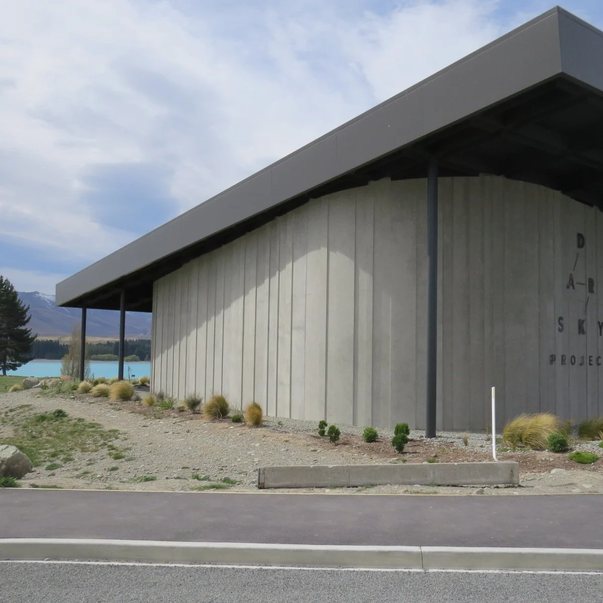 Modern building with a curved concrete wall and large overhanging roof, sign reads 'DARK SKY PROJECT', landscape includes mountains, trees, and a body of water in the background.
