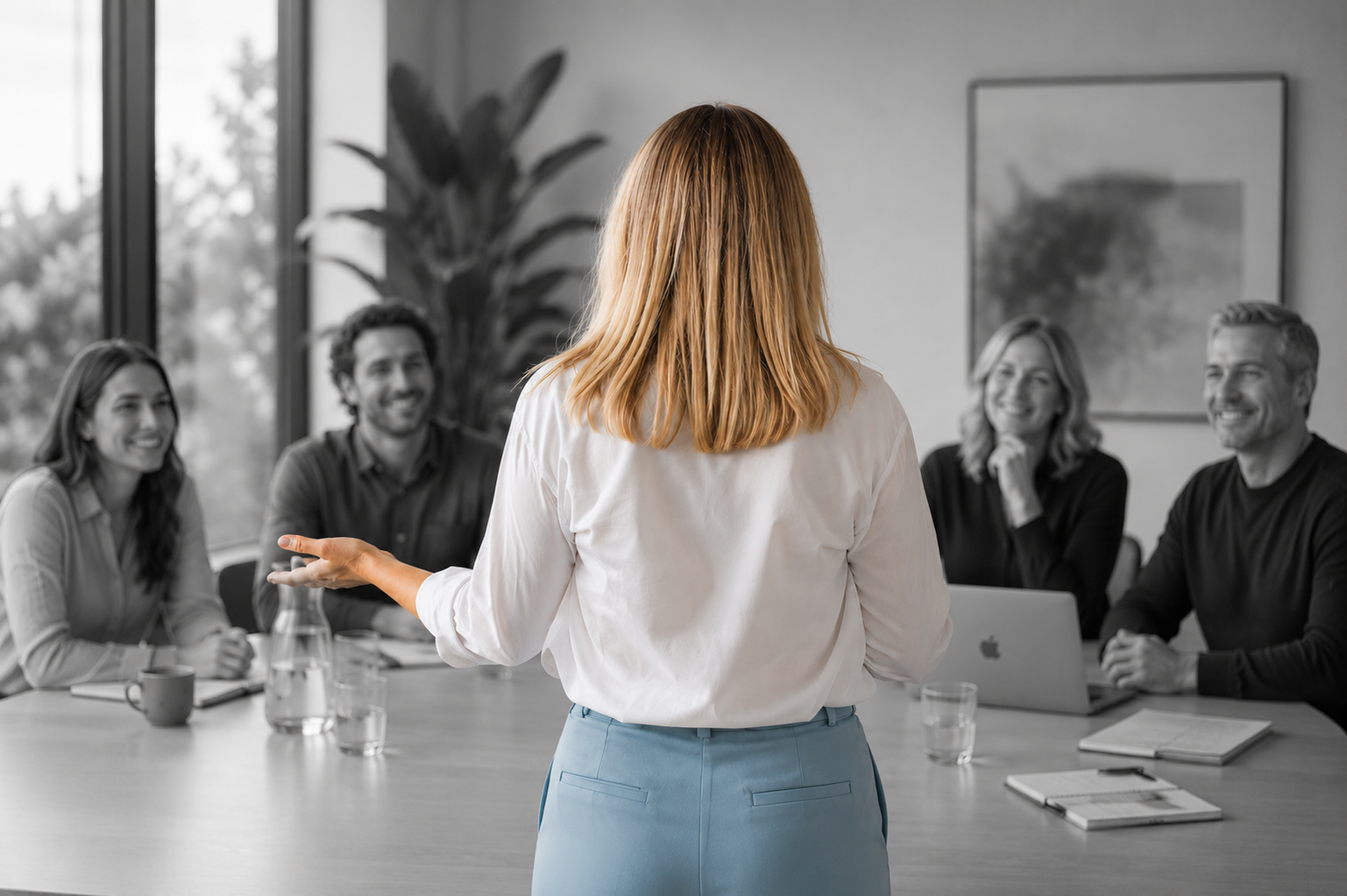 A woman giving a presentation to a diverse group of four people in a meeting room with large windows and a potted plant in the background.