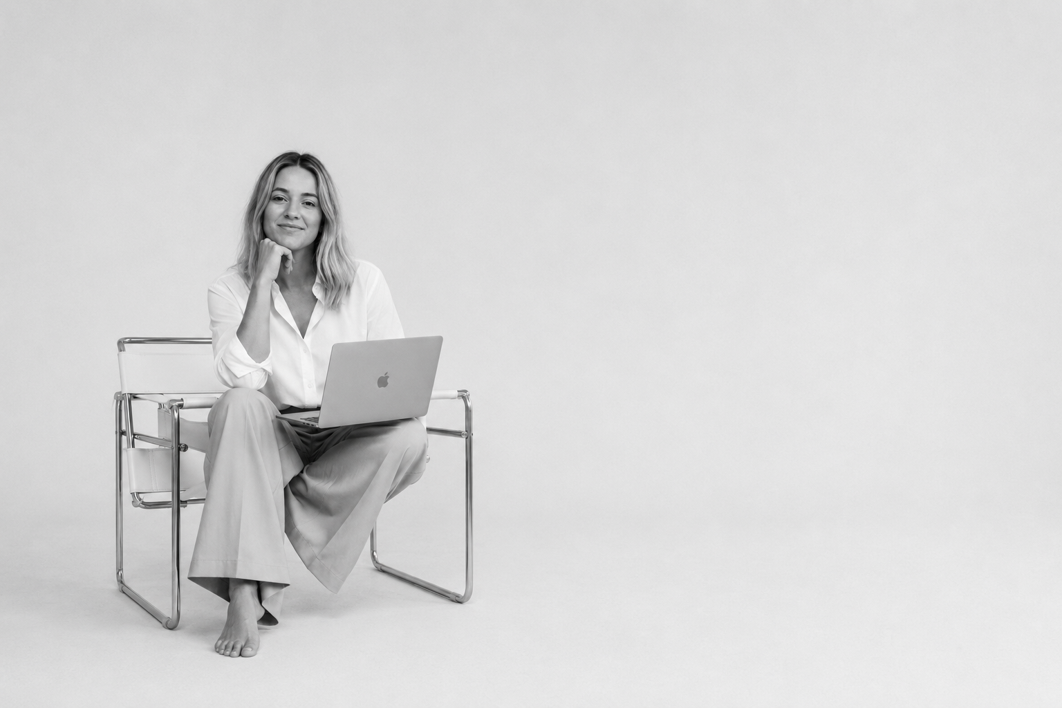 Black and white photo of a woman sitting barefoot on a modern metal chair, smiling, with a laptop on her lap, in an empty white room.
