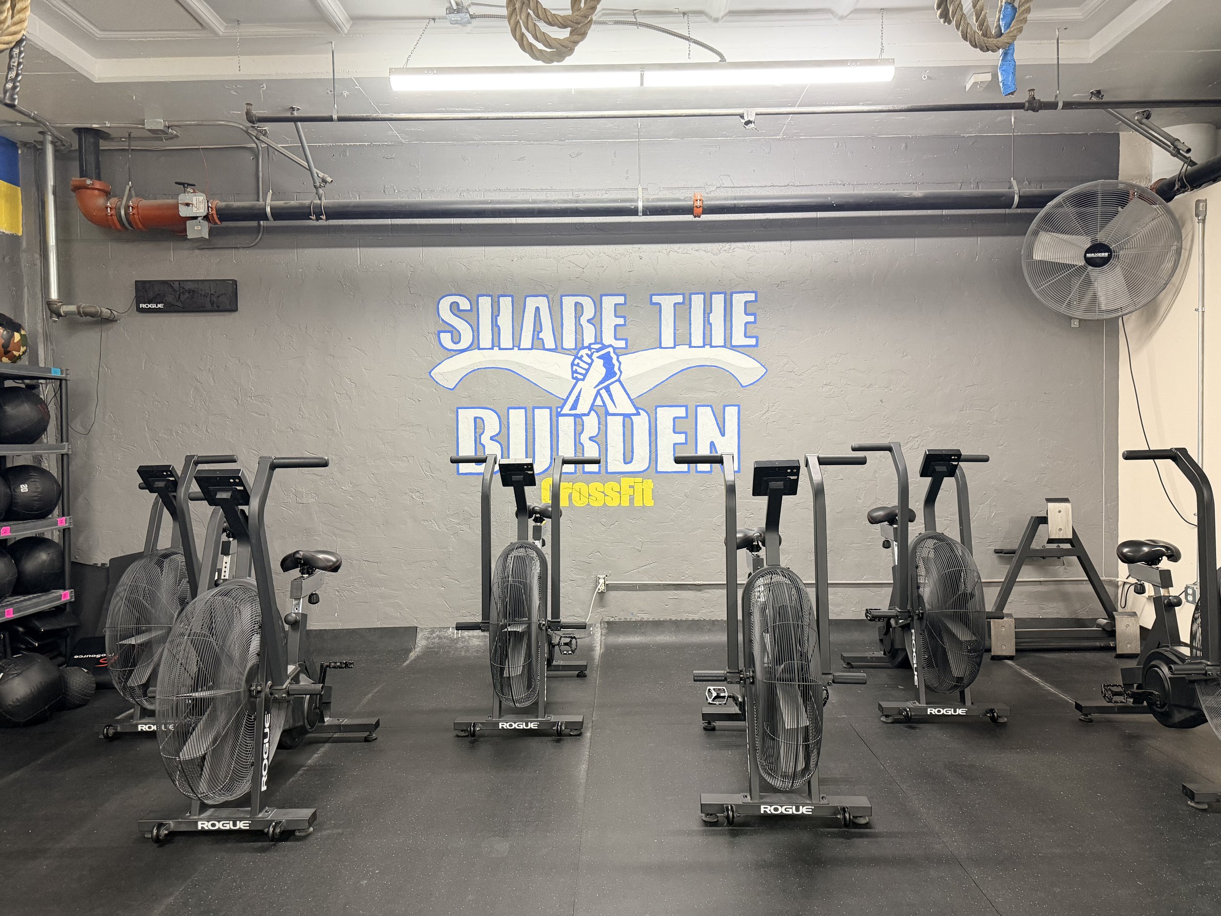 Indoor CrossFit gym with fan blades, rowing machines, and a mural on the wall that says "Share the Burn" with two fists bumping.