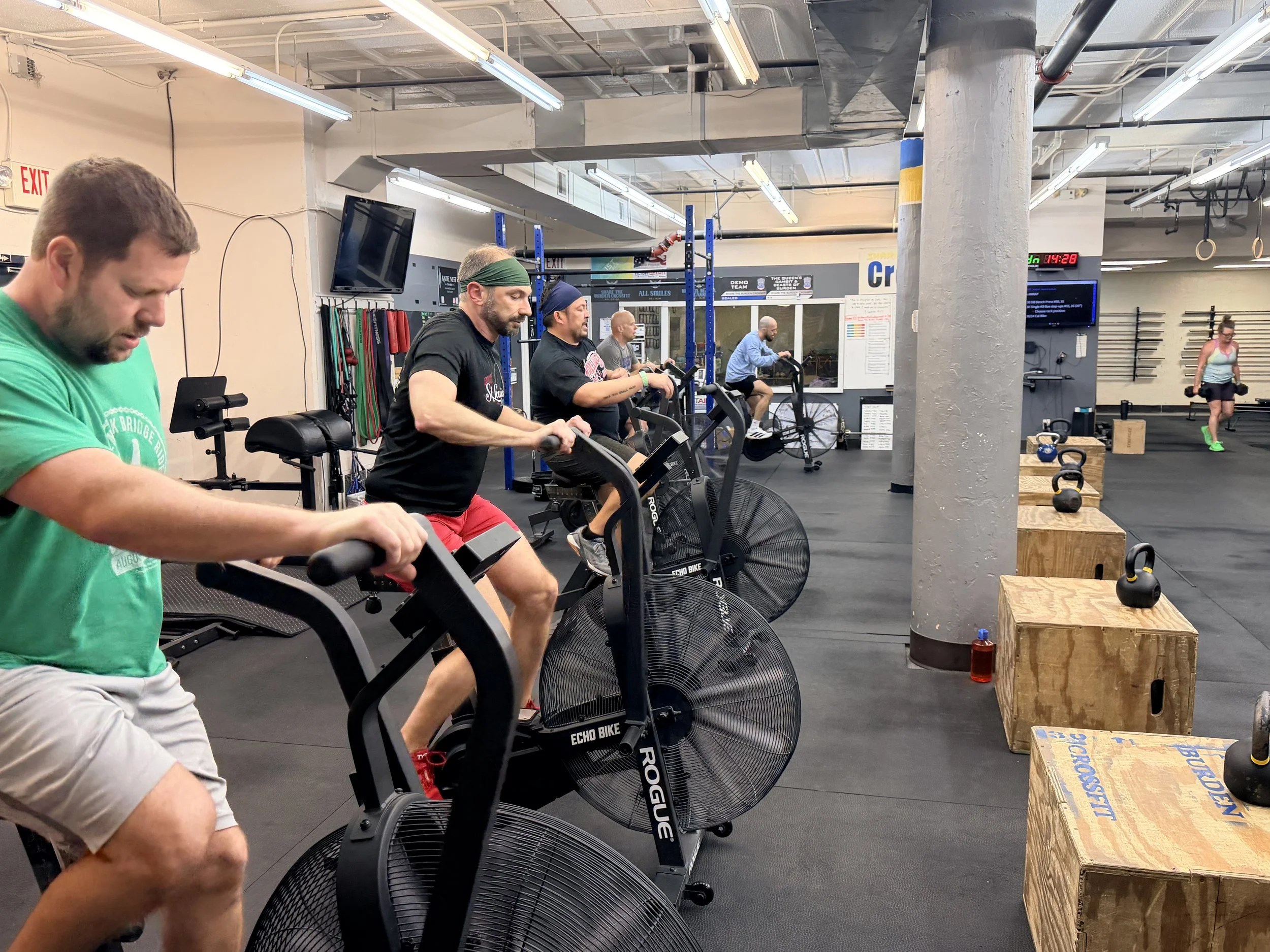 People exercising on stationary bikes in a gym with black floors, blue squat racks, hanging gym rings, and benches, with a woman walking in the background.