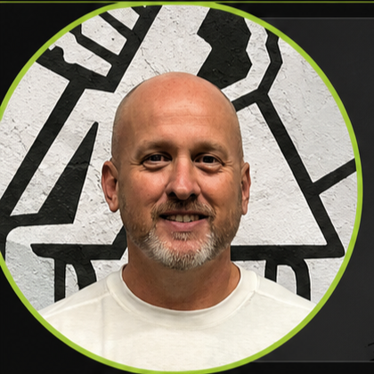 A smiling man with a shaved head and a beard, wearing a white shirt, standing in front of a black and white mural with abstract shapes.