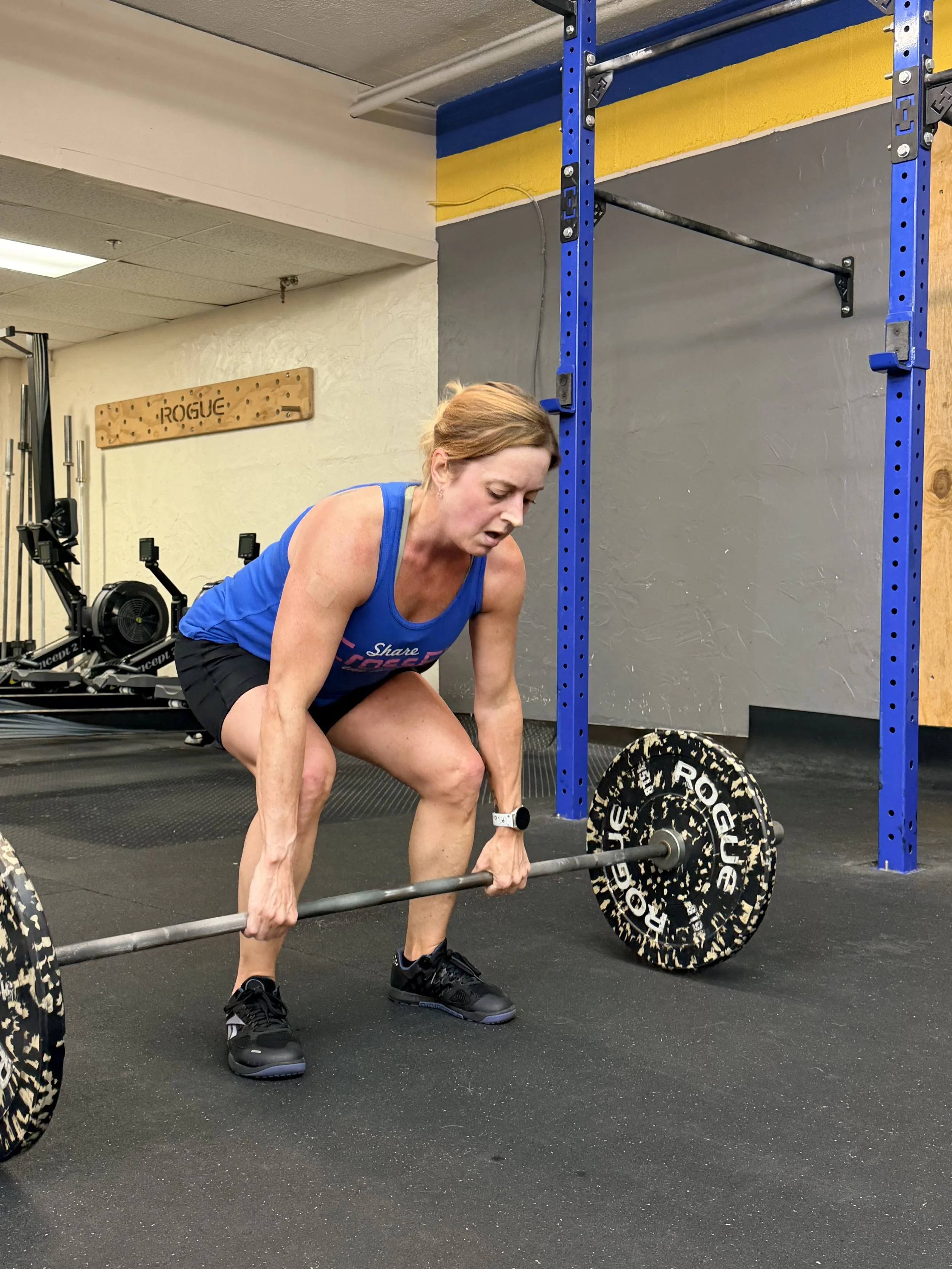 A woman with blonde hair tied back, wearing a blue tank top and black shorts, is performing a deadlift with a barbell loaded with Rogu weight plates in a gym. She is in a bent-over position, gripping the barbell with both hands, and is wearing black athletic shoes.