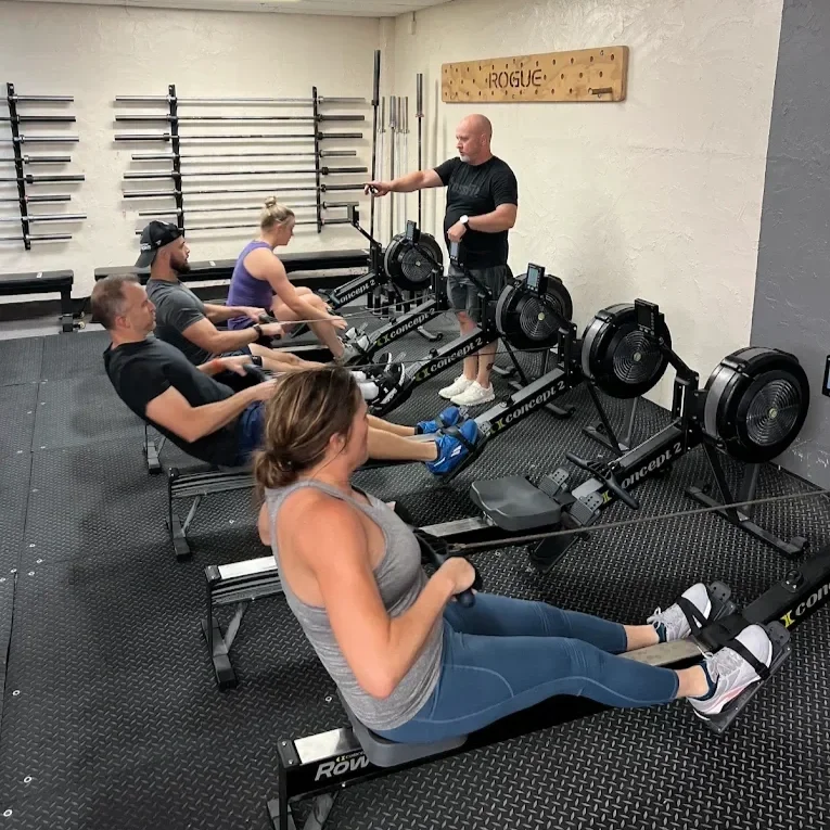 A fitness instructor in a black T-shirt and shorts giving rowing exercise instructions to four people in a gym, using rowing machines.