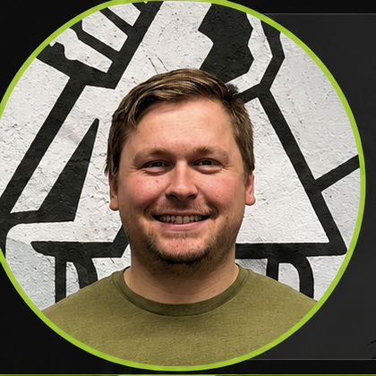 A smiling young man with short brown hair, wearing a green shirt, standing in front of a white wall with black geometric graffiti, bordered by a green circle.
