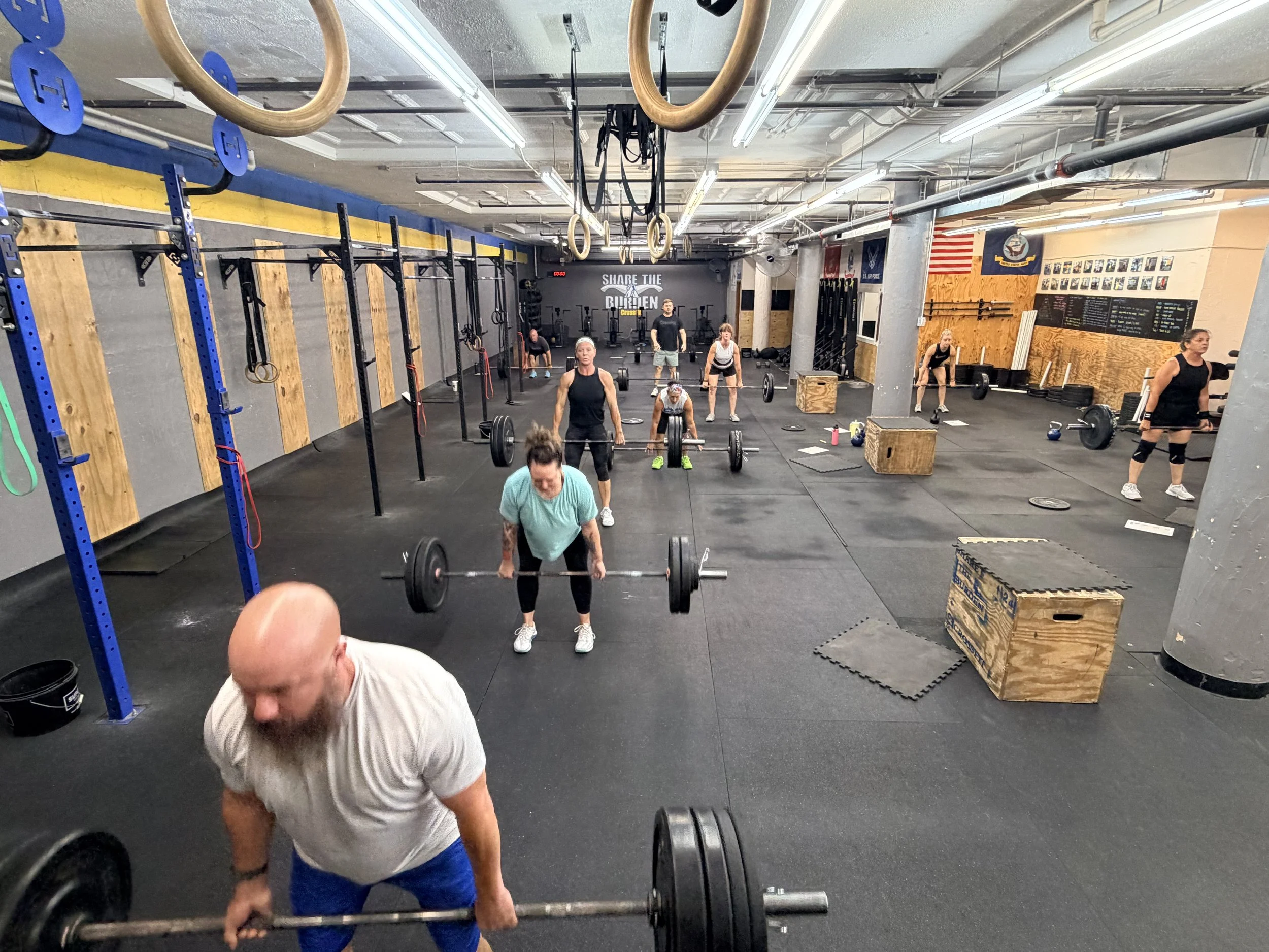 Indoor gym with people lifting weights and performing workouts, including deadlifts. Gym features black flooring, wooden wall panels, and gymnastics rings hanging from the ceiling. Multiple individuals are engaged in various exercises.