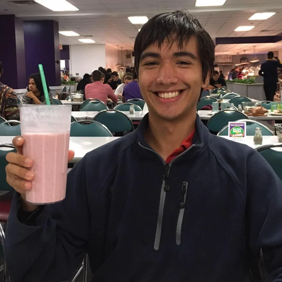 A young man smiling and holding a pink milkshake in a restaurant or cafeteria setting.