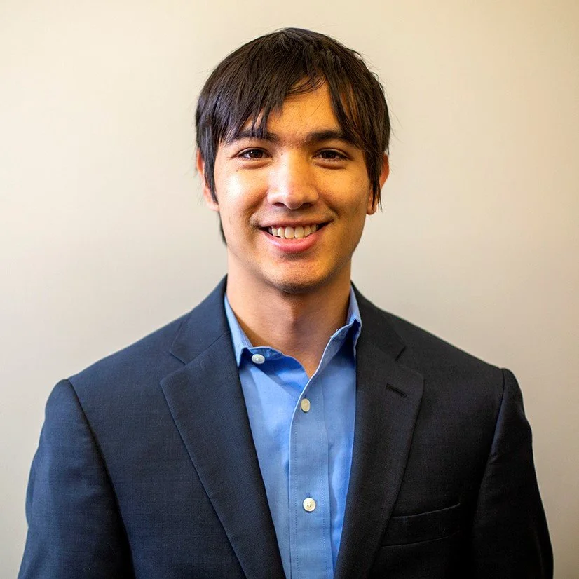 Young man with dark hair wearing a navy blazer and blue dress shirt, smiling against a plain wall.