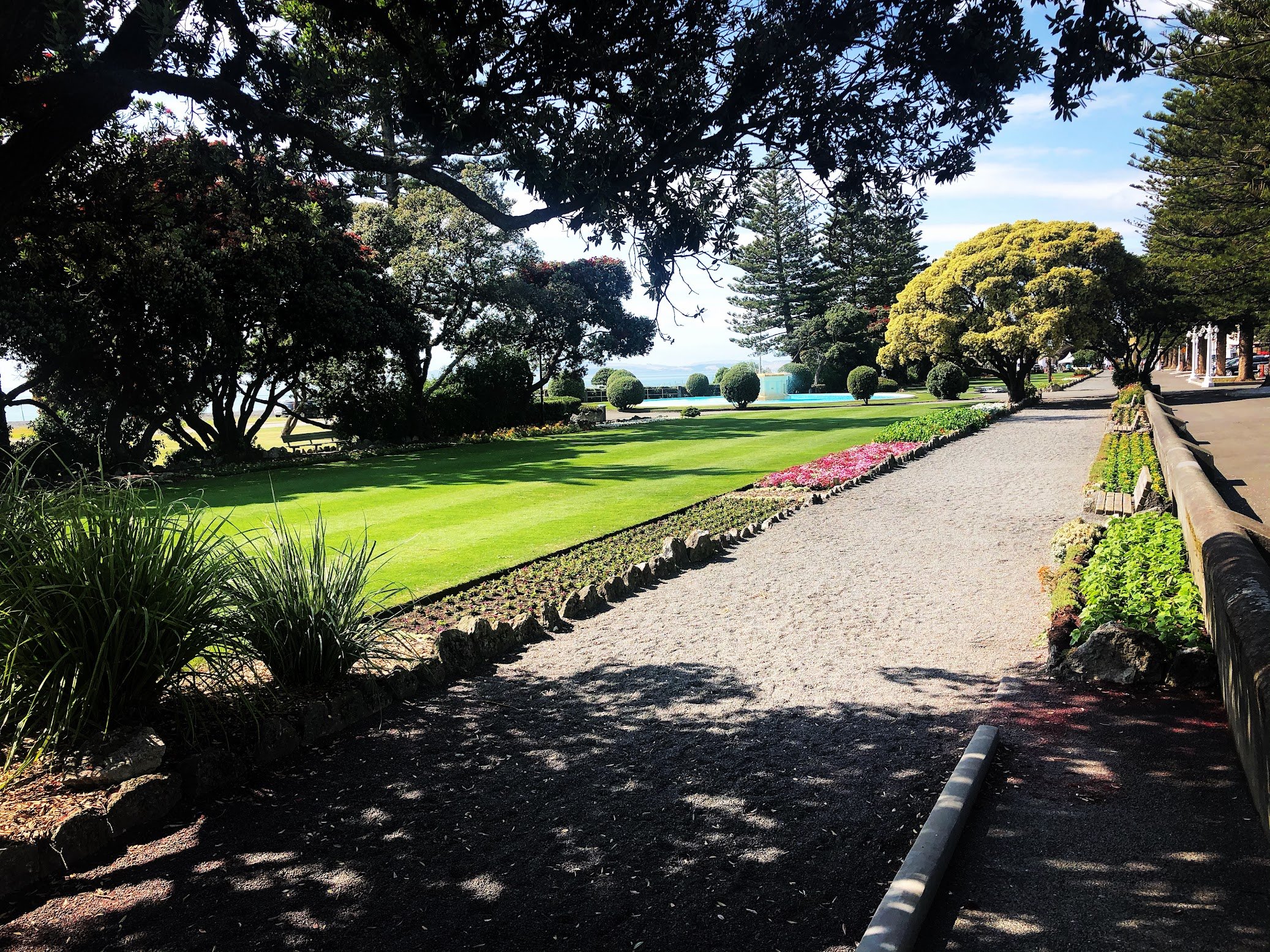 A park scene with sunlight filtering through trees, a well-maintained lawn, flowerbed, pathway, and a pool or pond in the background.