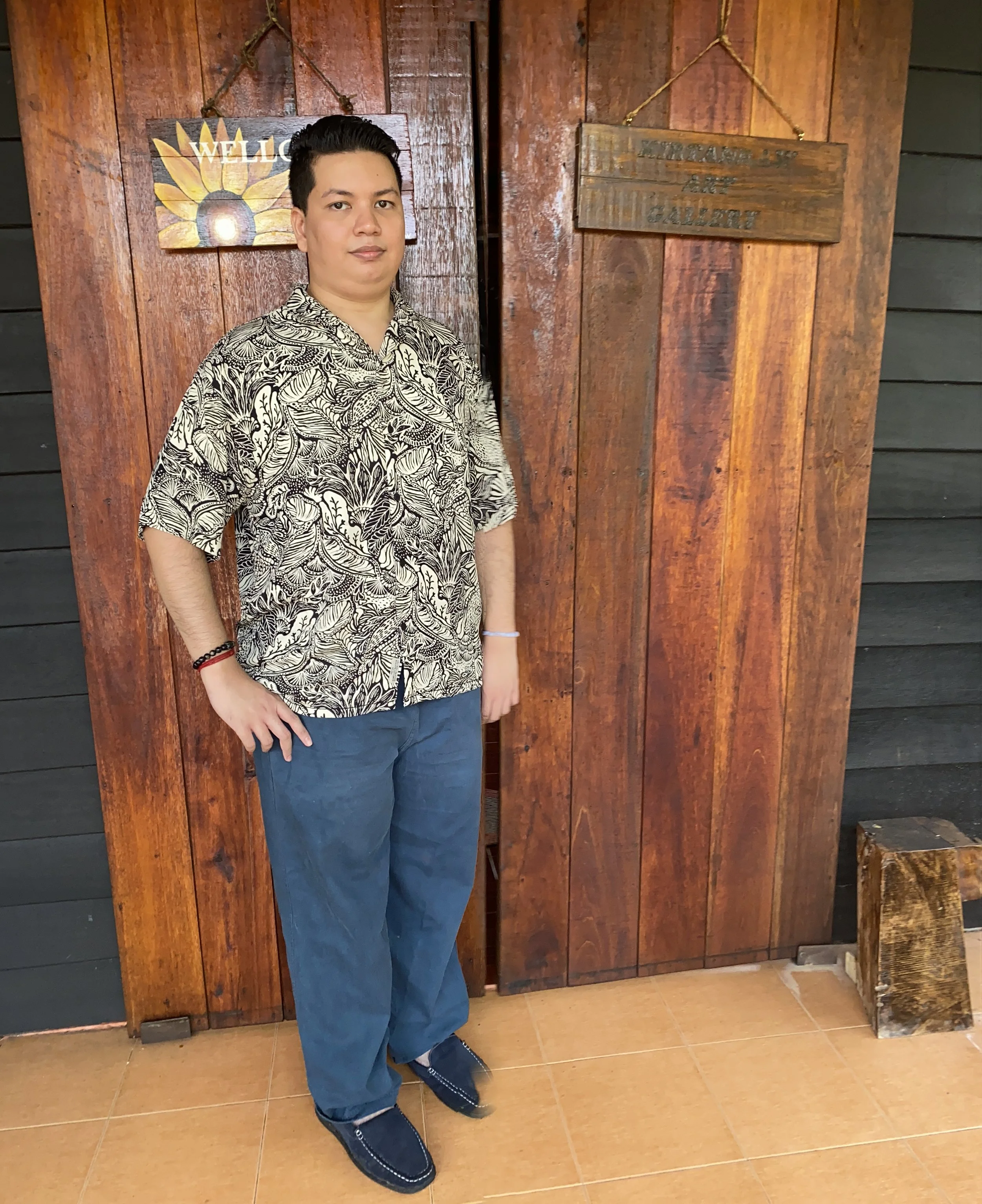 A young man standing in front of wooden wall panels with two decorative signs, one says 'Welcome' with a sunflower design, and the other is weathered and says 'Welcome to our cabin'. He is wearing a patterned shirt, blue pants, and black shoes with blue detailing, and has a focused expression.