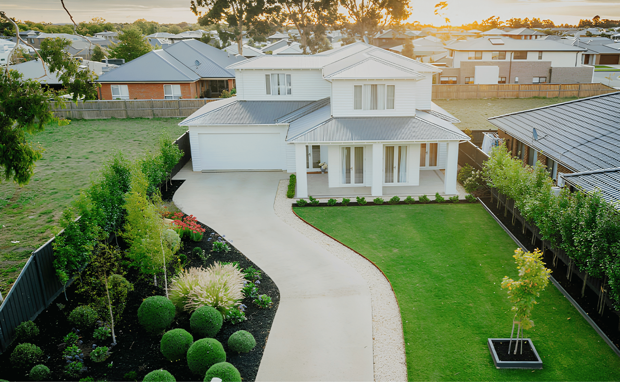 A modern two-story house with a metal roof, white siding, and large windows, surrounded by a well-maintained yard with flowerbeds, trees, and a curved concrete pathway at sunset.